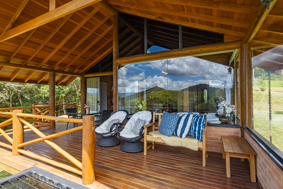 Modern wooden house on stilts with large glass windows, surrounded by grassy lawn and mountains under a clear blue sky.