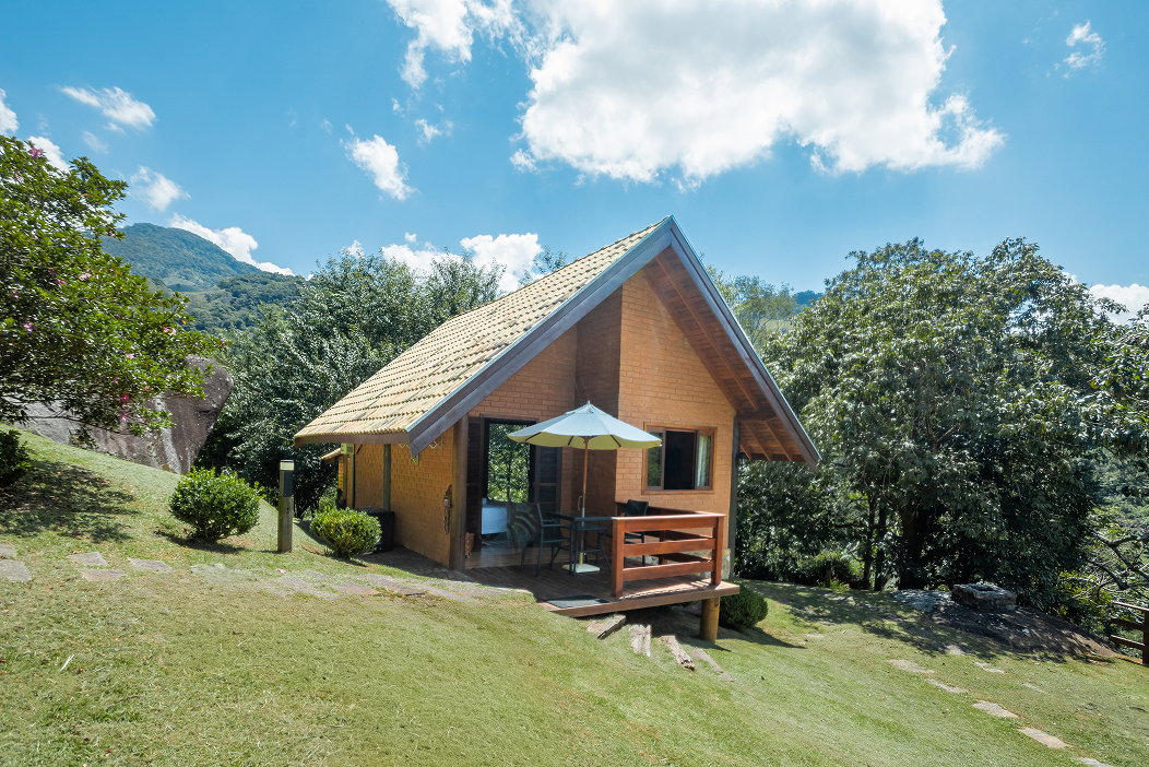 Wooden deck with black chair and table next to a cabin, overlooking a lush green mountainous landscape with clear sky and a large yellow plant in the foreground.