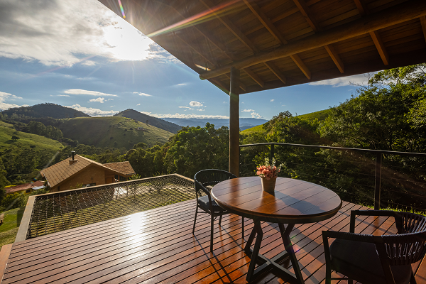 Wooden deck with black chair and table next to a cabin, overlooking a lush green mountainous landscape with clear sky and a large yellow plant in the foreground.